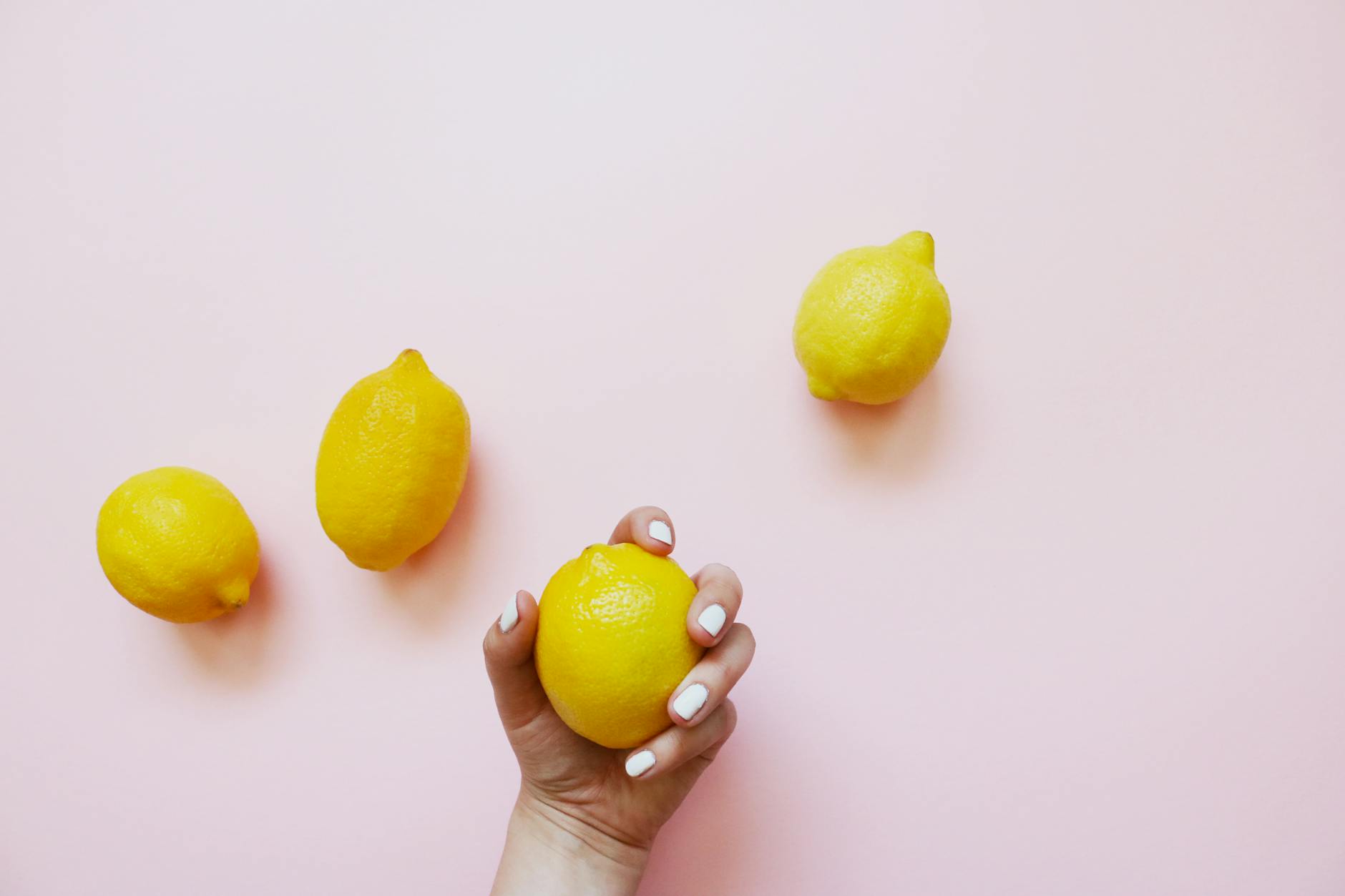 A hand with white nails holding a lemon on a soft pink background surrounded by additional lemons