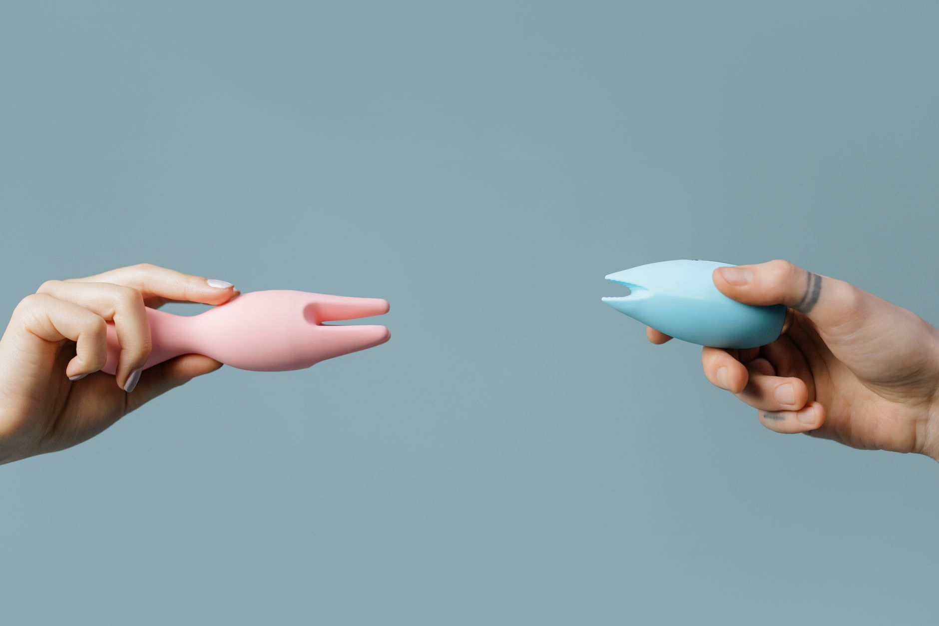 Two hands holding colorful silicone vibrators against a soft pastel background